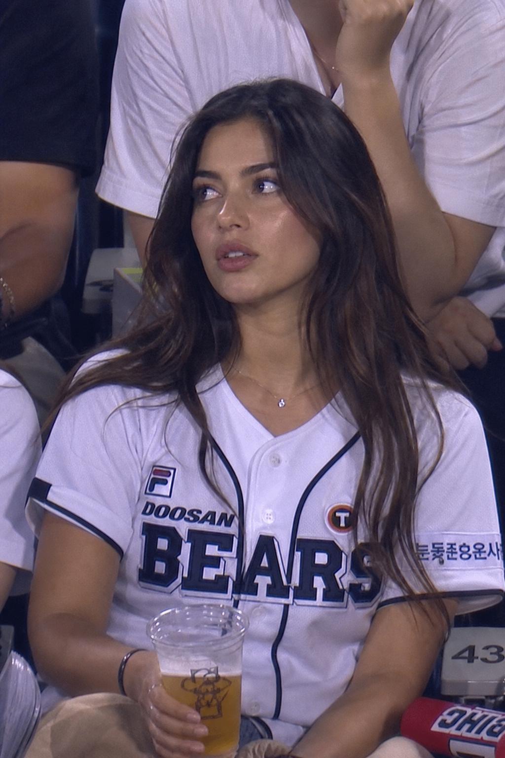 A Woman Wearing A Doosan Bears Jersey Enjoys A Drink While Sitting In A Stadium Surrounded By Other Fans