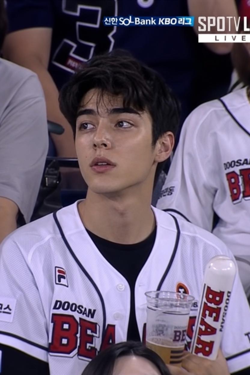 A Man Wearing A Doosan Bears Jersey Is Seated At A Baseball Game Holding A Drink And A Team-Branded Inflatable Cheering Stick