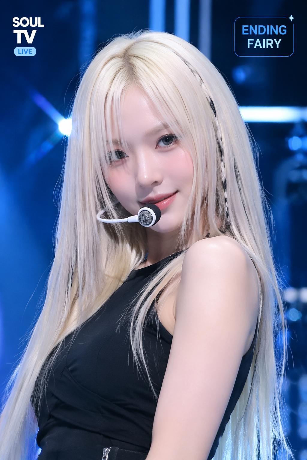 This image shows a young female idol performing on a Korean music show stage. She has lon...