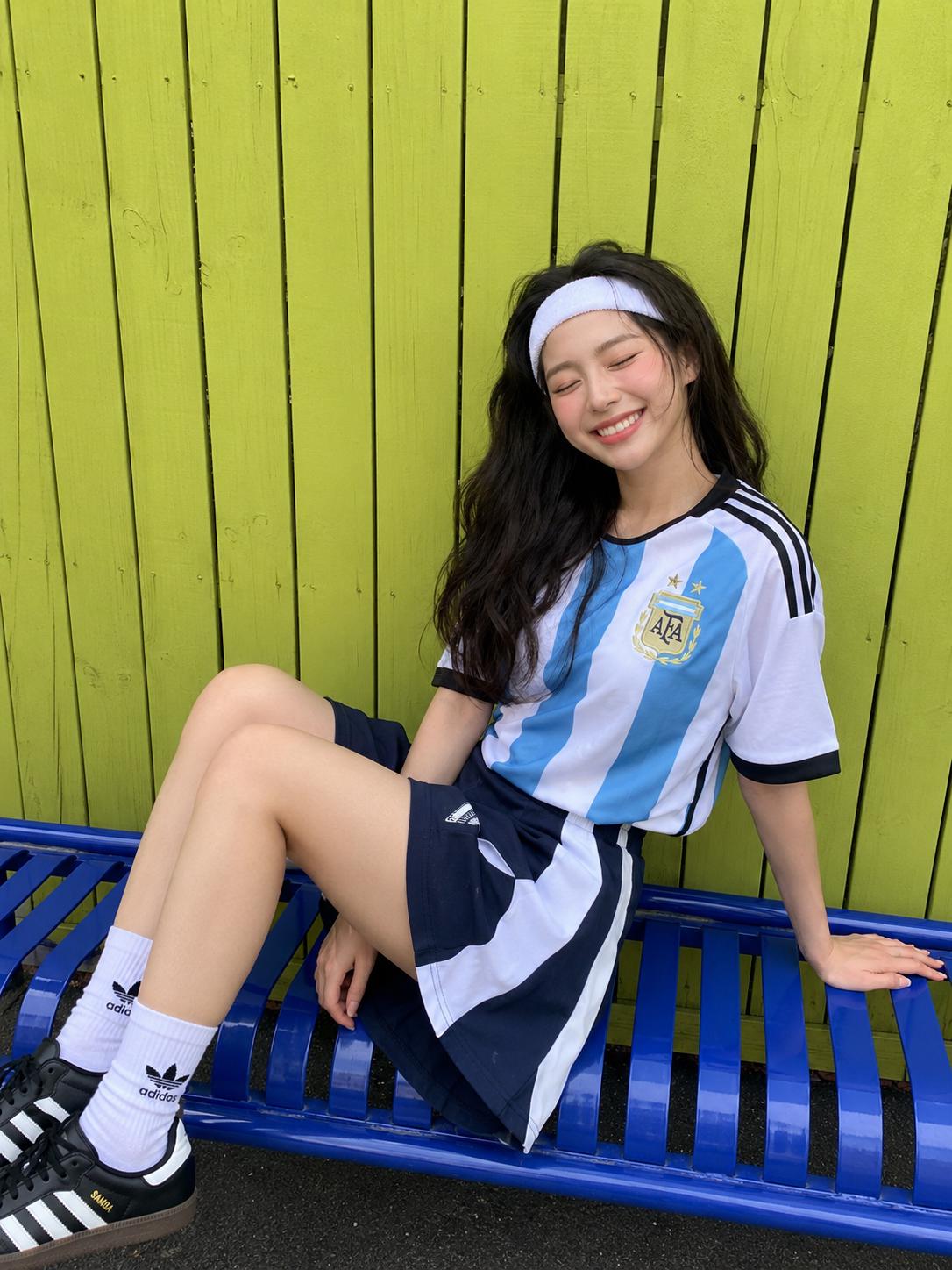 A Woman Is Sitting On A Blue Bench Against A Bright Green Wooden Fence Wearing A Sports Outfit