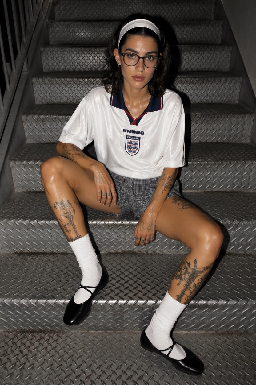 A Woman Wearing An England Soccer Jersey And Patterned Shorts Sits On Metal Stairs In An Urban Setting