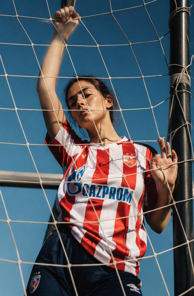A Woman In A Striped Soccer Jersey Stands Behind A Net Under A Clear Blue Sky