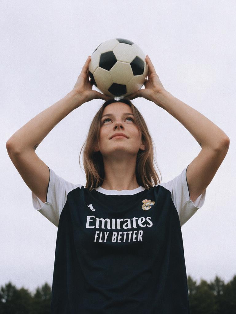A Woman In A Soccer Jersey Is Holding A Ball Above Her Head Outdoors With A Cloudy Sky And Trees In The Background