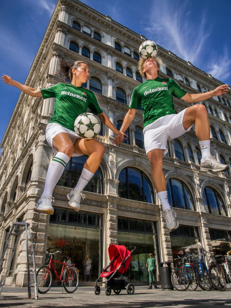 Two People Wearing Green Jerseys Perform A Synchronized Soccer Trick In Front Of A Historic Building On A Sunny Urban Street