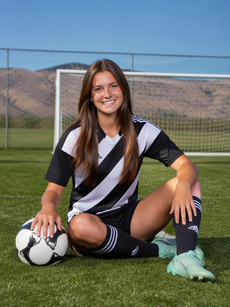 A Woman In A Soccer Uniform Is Sitting On A Field With A Ball In Front Of A Goal And Mountain Backdrop