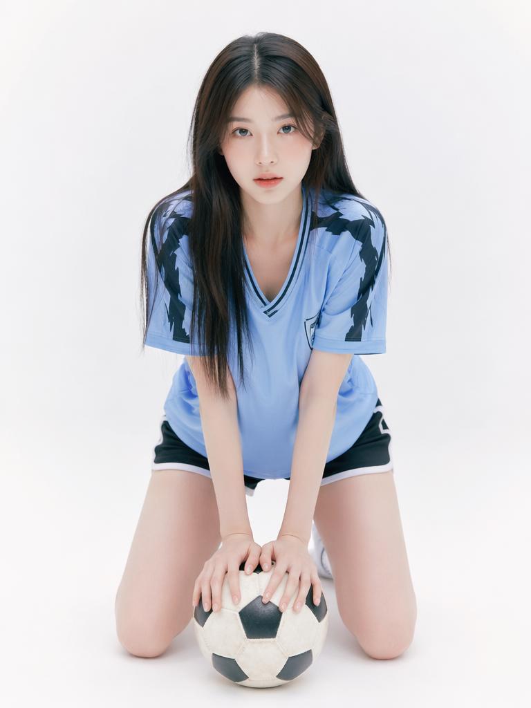 A Woman In A Blue Soccer Jersey Kneels On A White Background While Holding A Soccer Ball