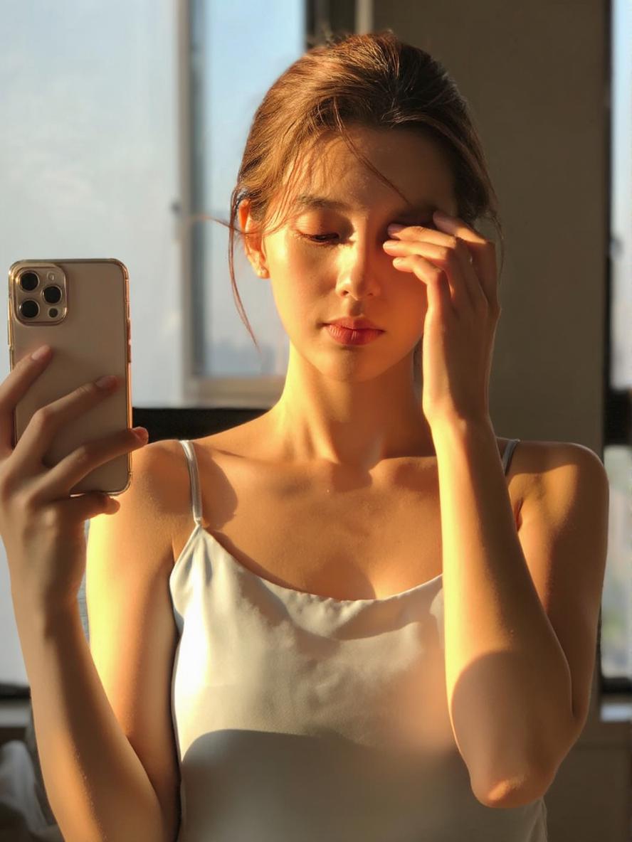 A Woman Stands Near A Window In Soft Sunlight Holding A Smartphone And Capturing A Reflective Moment